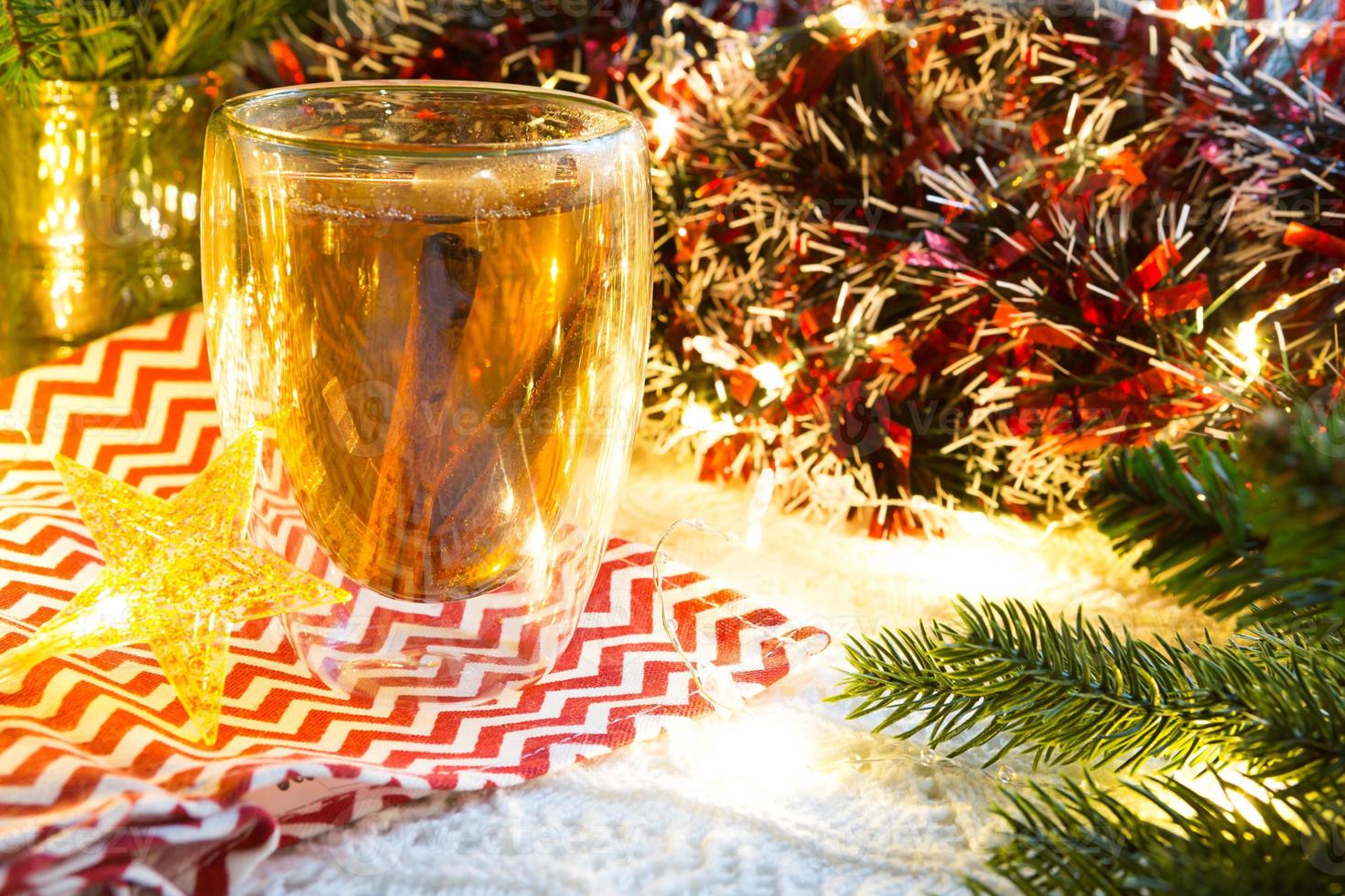 transparenter doppelwandiger glasbecher mit heißem tee und zimtstangen auf dem tisch mit weihnachtsdekor. Silvester-Atmosphäre, getrocknete Orangenscheibe, Girlande und Lametta, Fichtenzweig, gemütlich foto