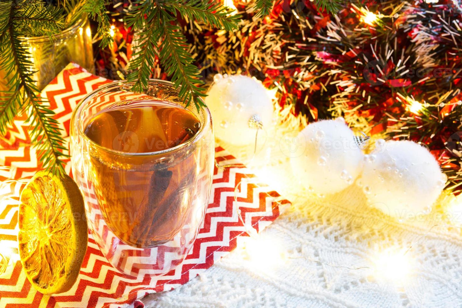 transparenter doppelwandiger glasbecher mit heißem tee und zimtstangen auf dem tisch mit weihnachtsdekor. Silvester-Atmosphäre, getrocknete Orangenscheibe, Girlande und Lametta, Fichtenzweig, gemütlich foto