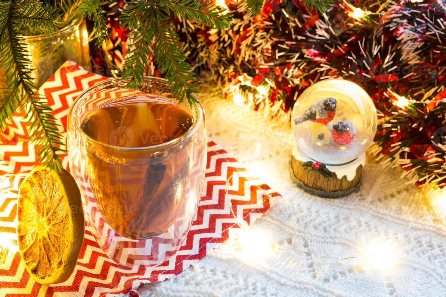 transparenter doppelwandiger glasbecher mit heißem tee und zimtstangen auf tisch mit weihnachtsdekor. Silvesterstimmung, getrocknete Orangenscheibe, Girlande und Lametta, Schneekugel mit Dompfaffen foto