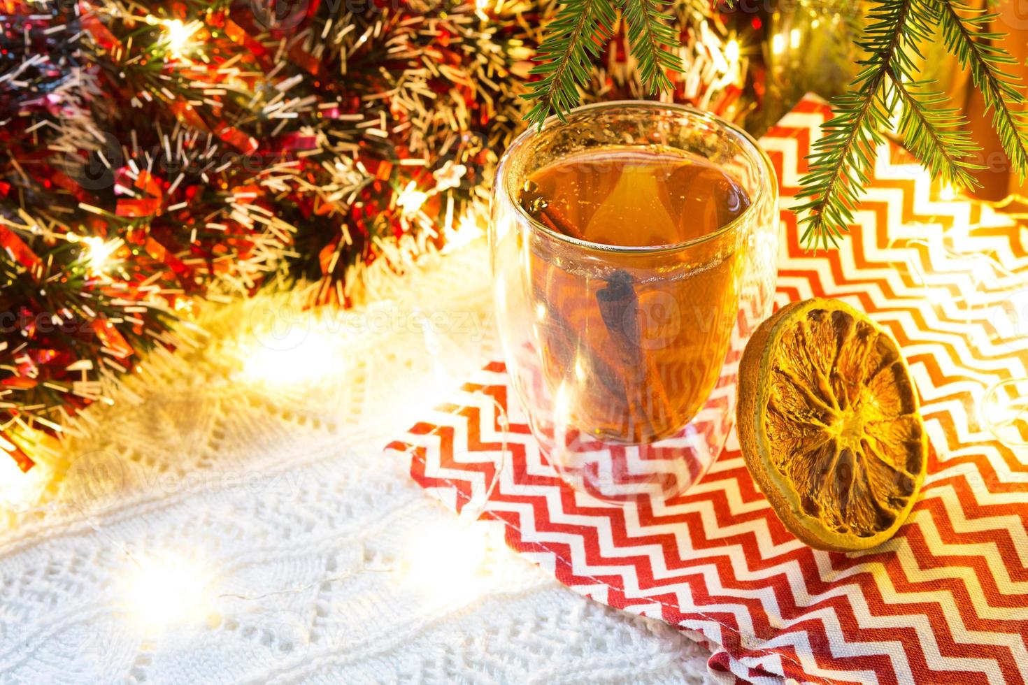 transparenter doppelwandiger glasbecher mit heißem tee und zimtstangen auf dem tisch mit weihnachtsdekor. Silvester-Atmosphäre, getrocknete Orangenscheibe, Girlande und Lametta, Fichtenzweig, gemütlich foto