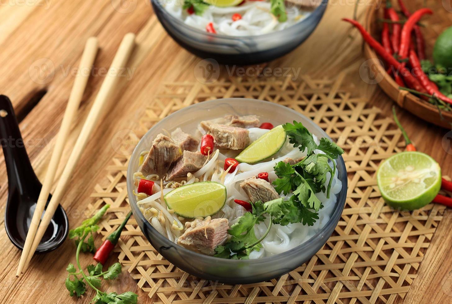 vietnam pho bo suppe mit rindfleisch, reisnudeln, limette, chilipfeffer in grauer schüssel auf holzhintergrund. foto
