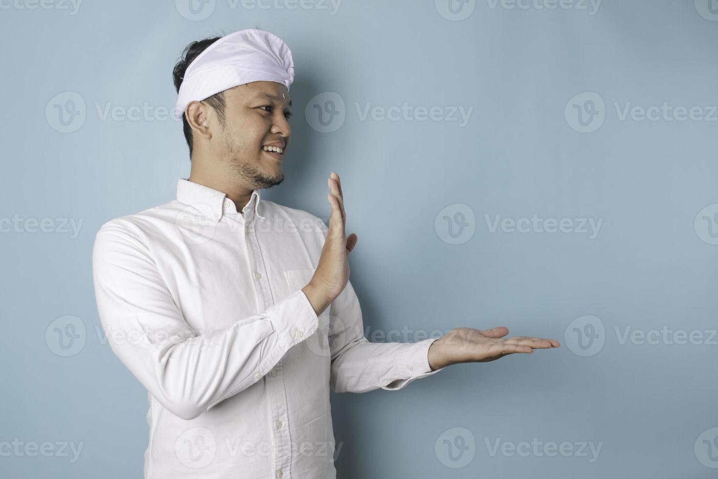 Aufgeregter balinesischer Mann mit Udeng oder traditionellem Stirnband und weißem Hemd, der auf den Kopierraum neben ihm zeigt, isoliert durch blauen Hintergrund foto