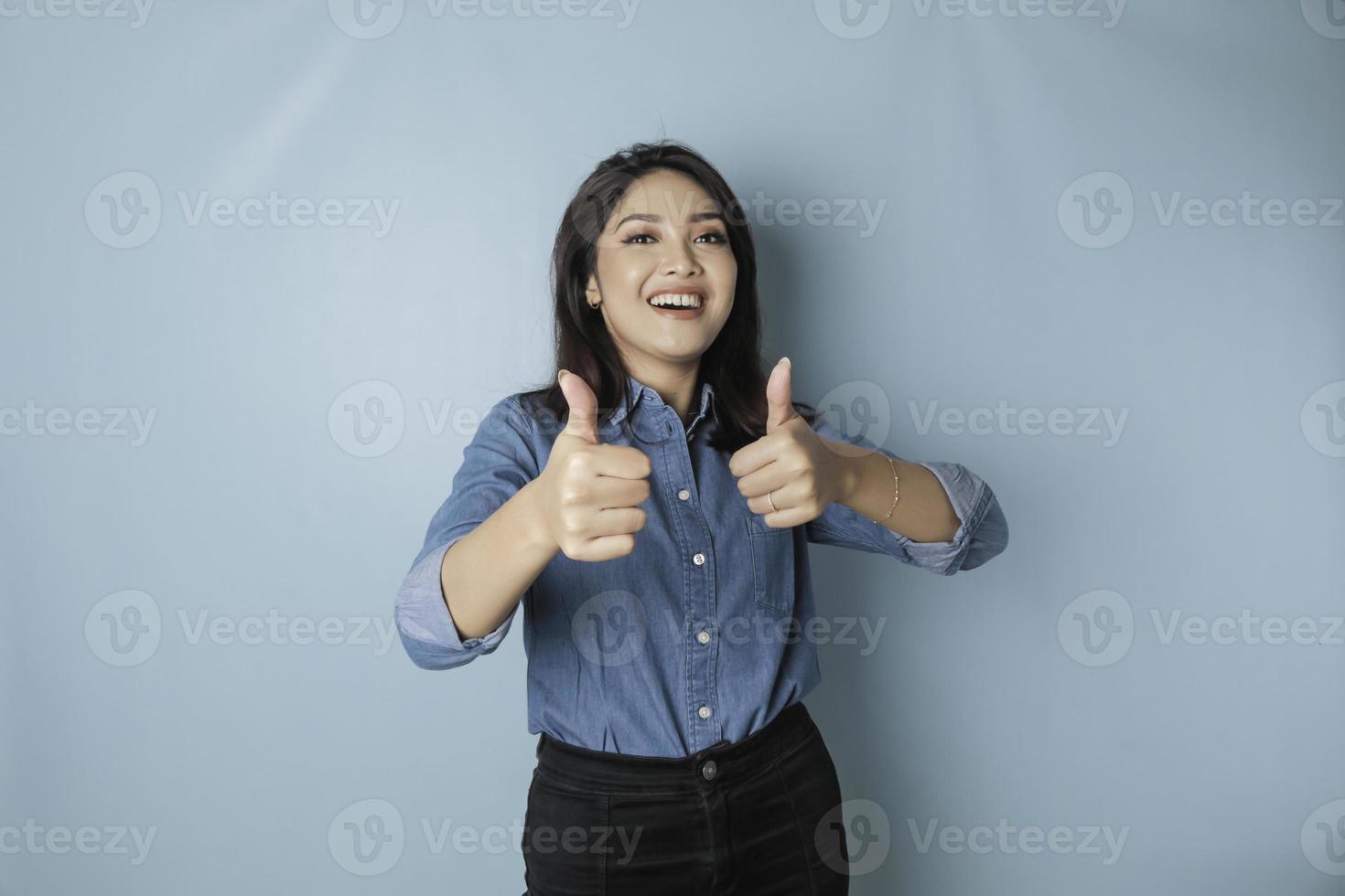 Aufgeregte asiatische Frau, die ein blaues Hemd trägt, gibt Daumen nach oben Handgeste der Zustimmung, isoliert durch blauen Hintergrund foto