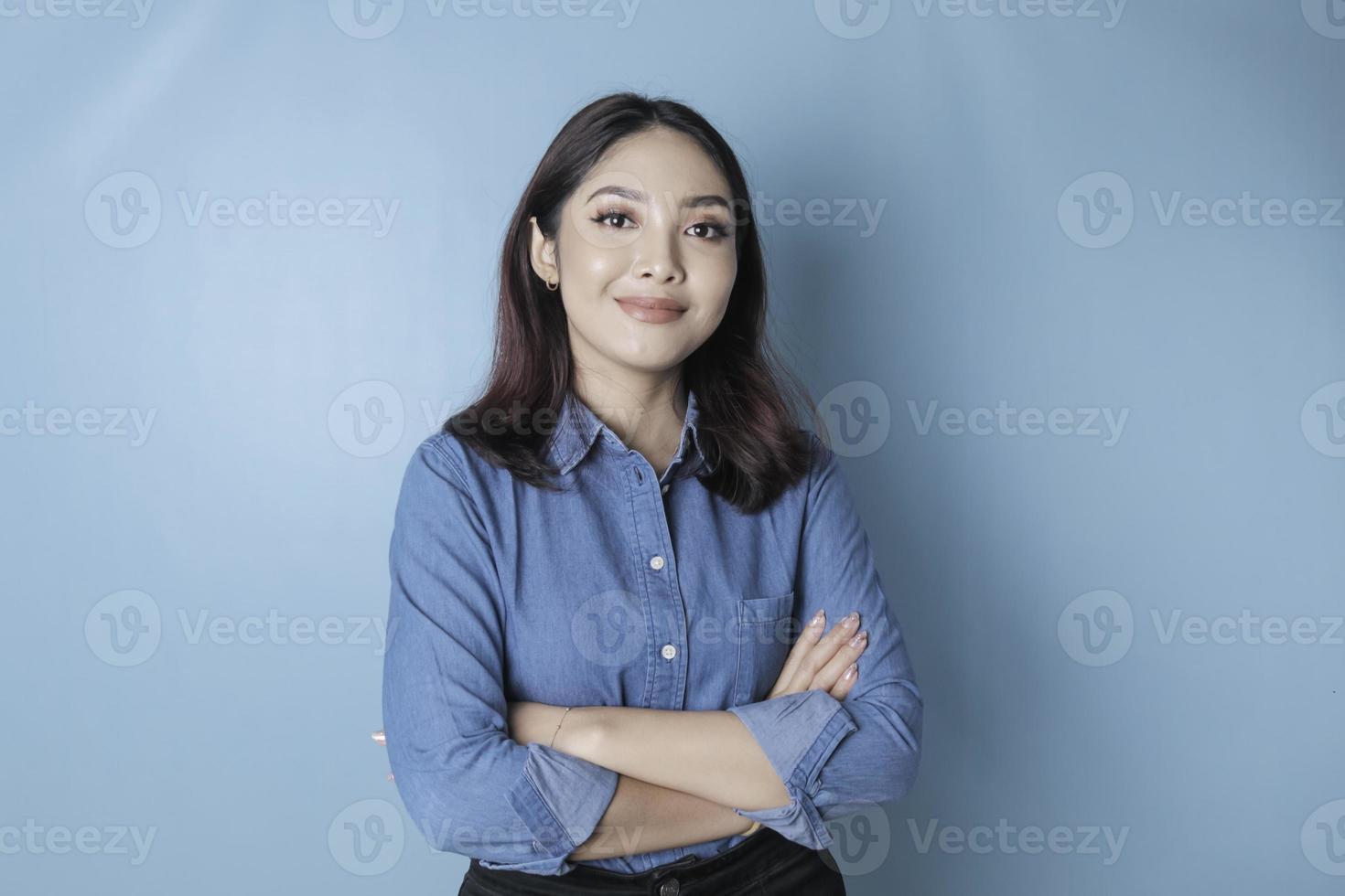 Porträt eines selbstbewussten lächelnden Mädchens, das mit verschränkten Armen steht und isoliert auf blauem Hintergrund in die Kamera schaut und ein blaues Hemd trägt foto
