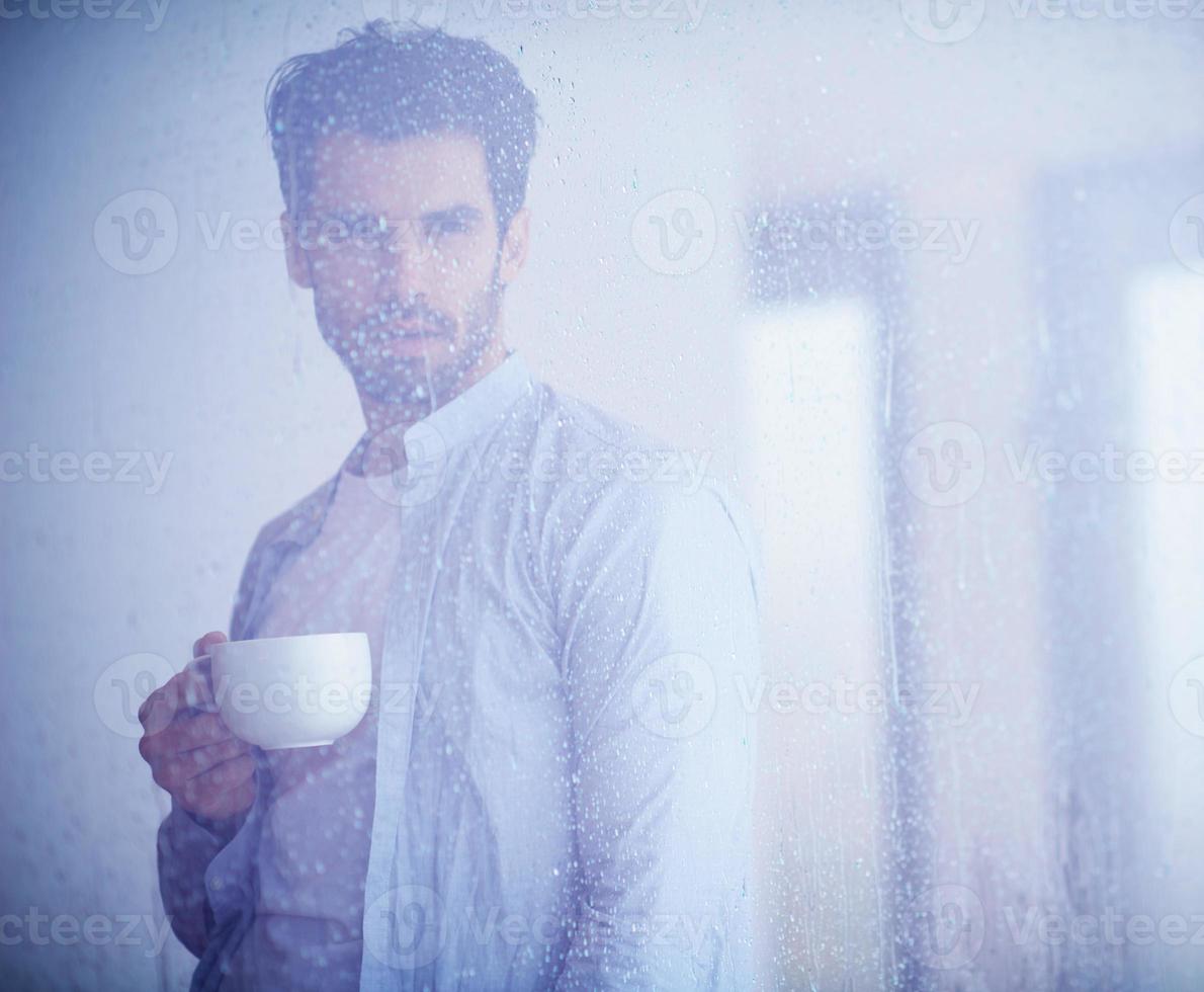 Entspannter junger Mann trinkt am ersten Morgen Kaffee mit Regentropfen auf dem Fenster foto