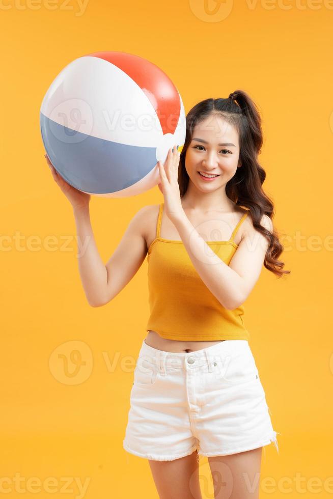 Lächelndes hübsches asiatisches Mädchen im Sommeroutfit mit buntem Wasserball auf isoliertem gelbem Hintergrund foto