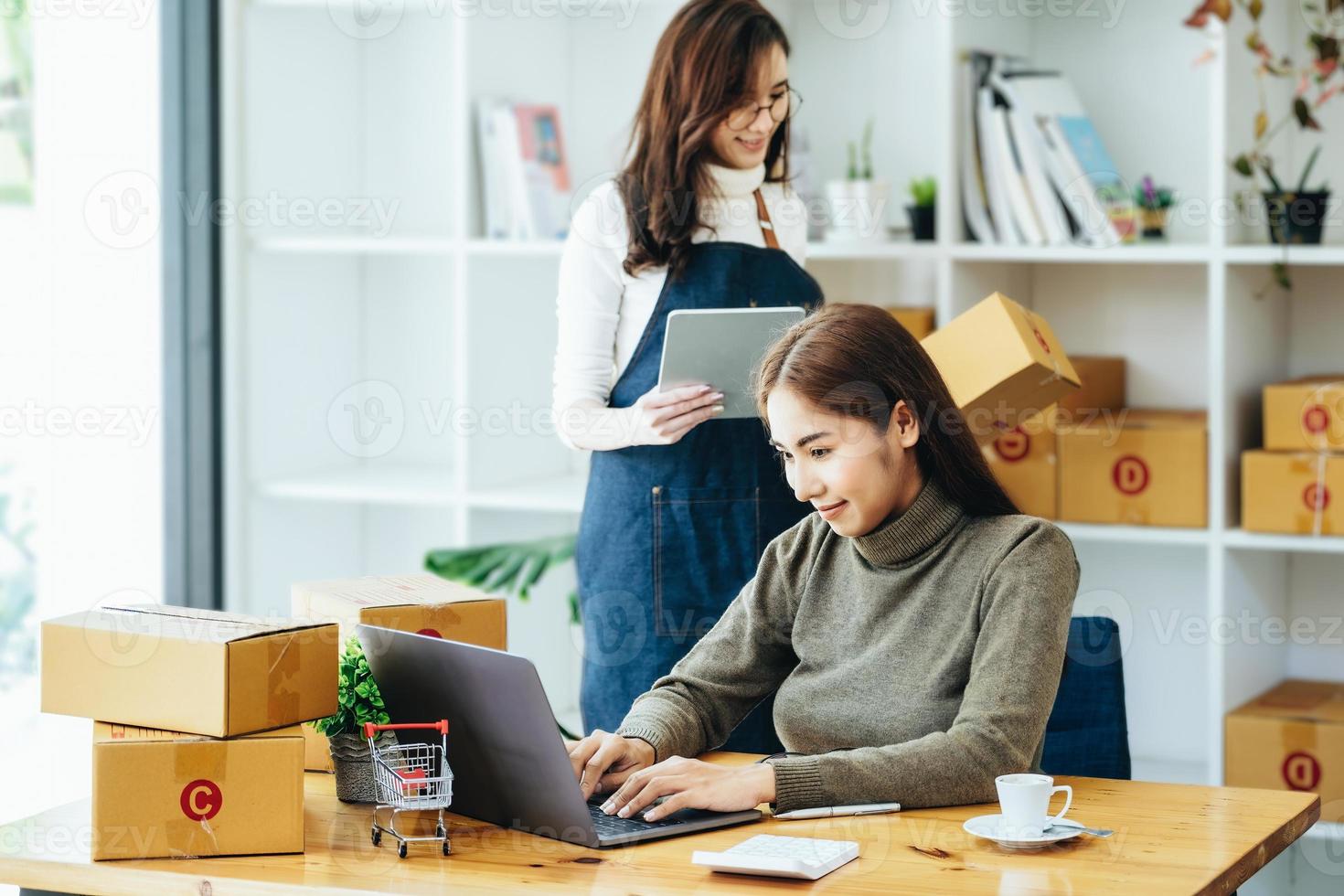 Zwei glückliche Frauen, die Produkte online verkaufen, gründen einen Kleinunternehmer, der einen Laptop und ein Tablet verwendet, um Preise zu berechnen und den Versand vorzubereiten. foto