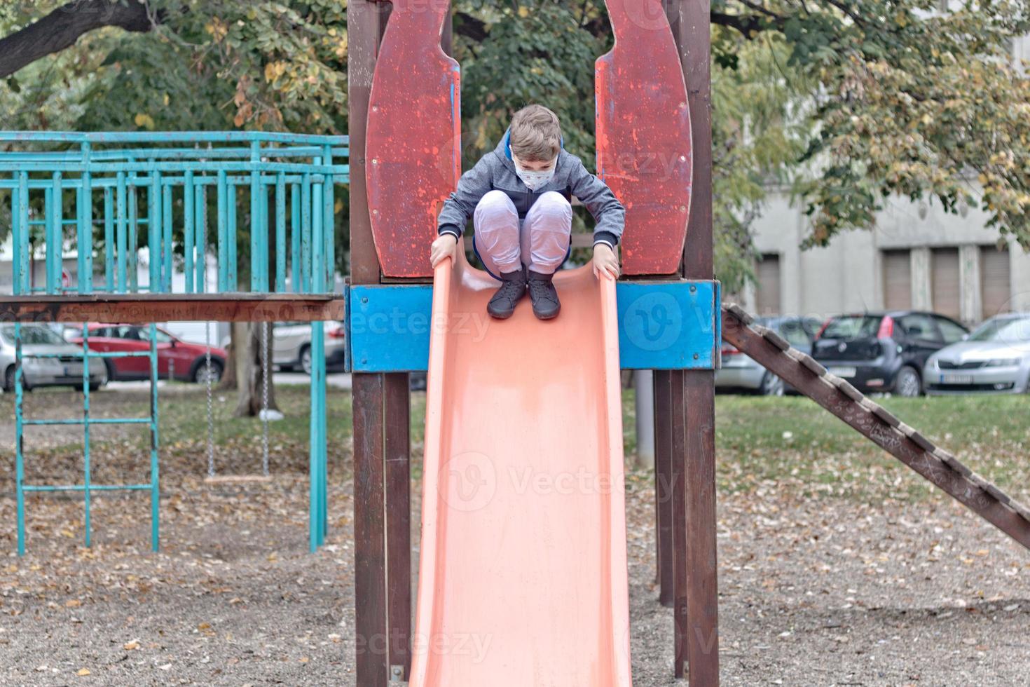 kleiner Junge mit Gesichtsmaske beim Rutschen im Park. foto