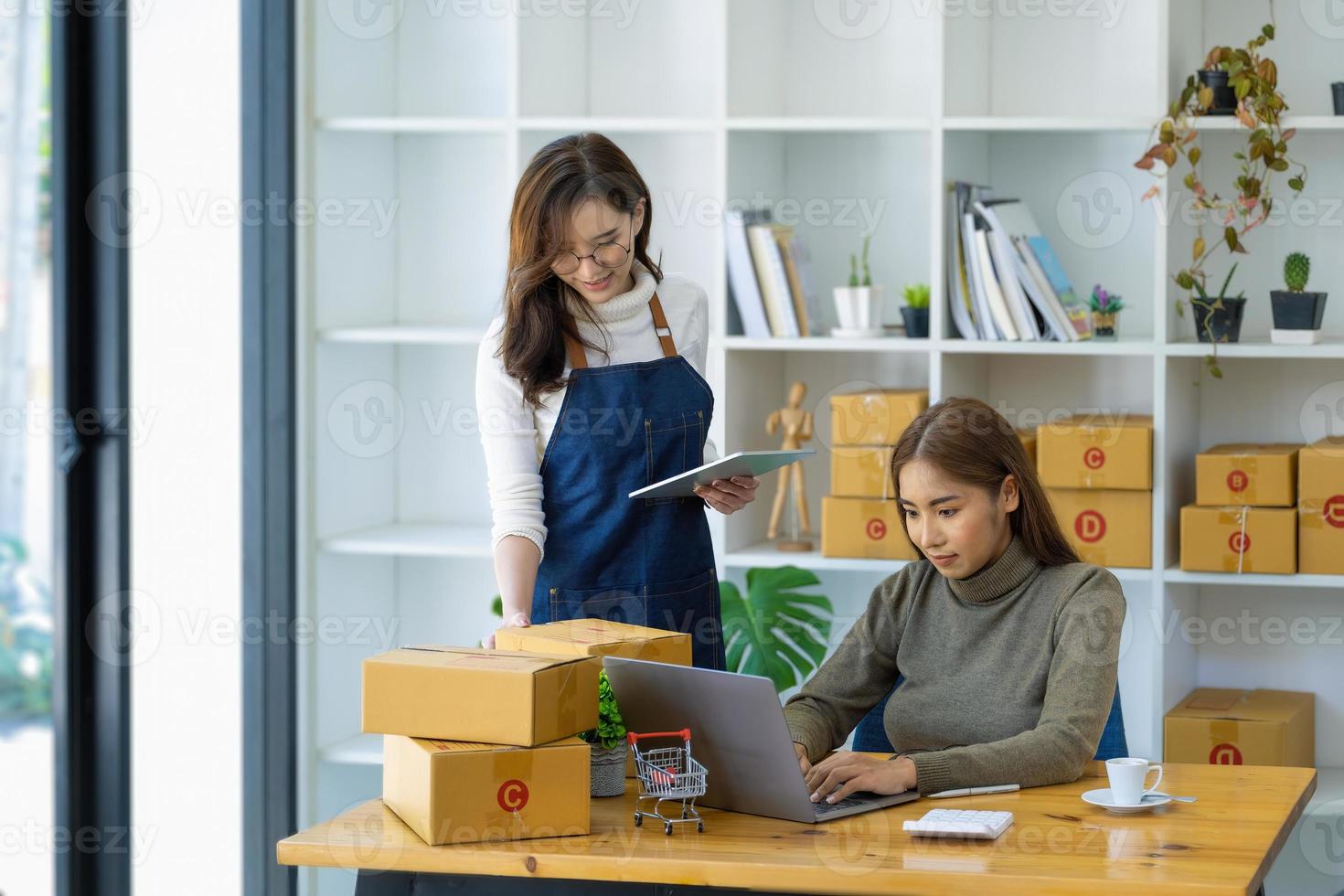 Zwei glückliche Frauen, die Produkte online verkaufen, gründen einen Kleinunternehmer, der einen Laptop und ein Tablet verwendet, um Preise zu berechnen und den Versand vorzubereiten. foto