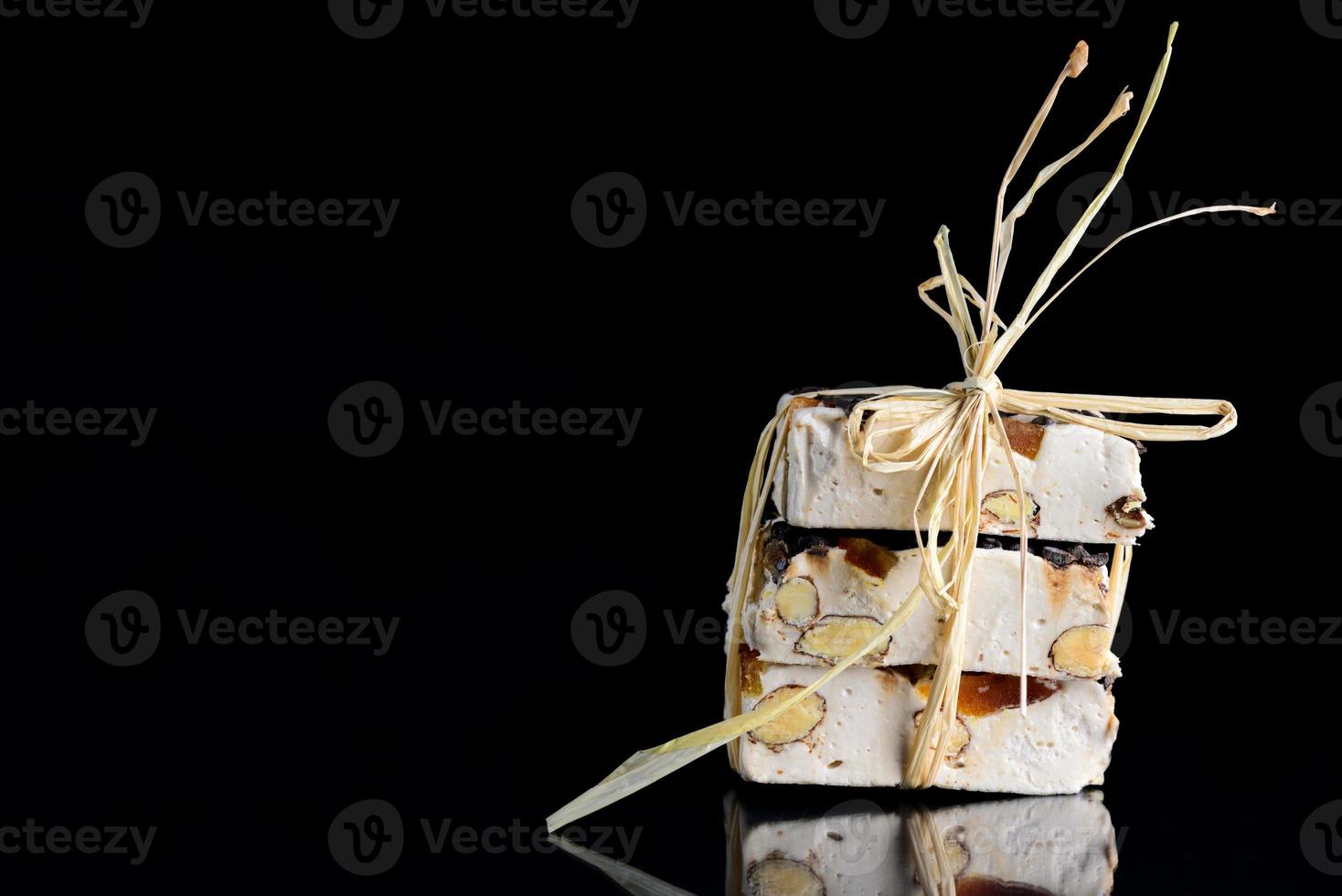 mehrere torrone- oder weiße nougatstücke mit nüssen, kandierten früchten und schokolade sind mit bast zusammengebunden und liegen auf einer spiegelnden fläche vor dunklem hintergrund foto