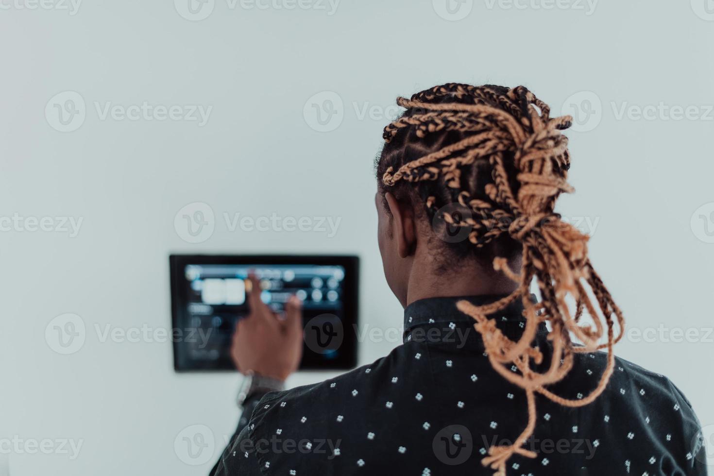 Lächelnder afroamerikanischer Mann mit modernem Smart-Home-System, Controller an der Wand, positiver junger Mann, der die Temperatur am Thermostat schaltet oder Sicherheitsalarm in der Wohnung aktiviert foto