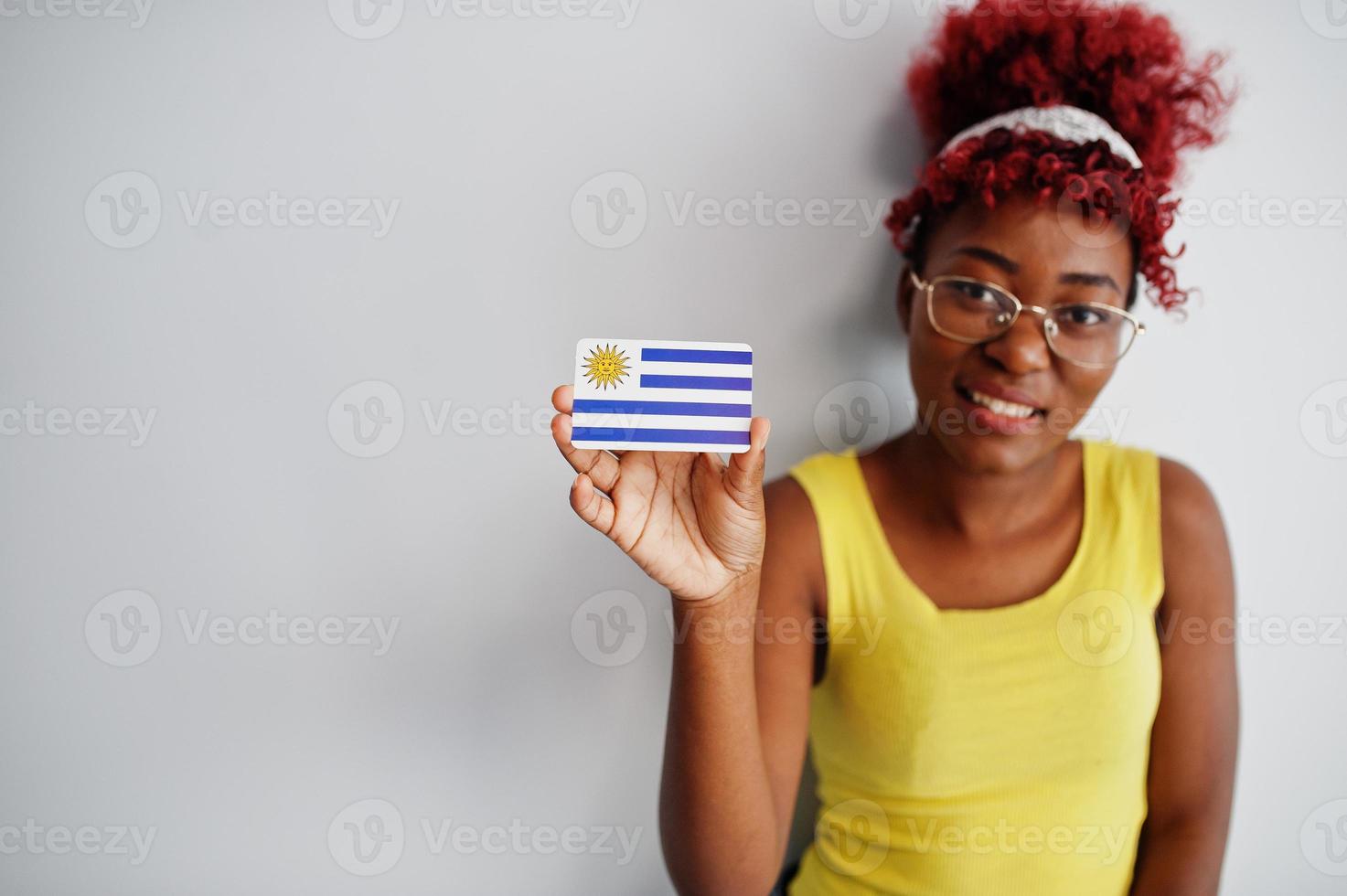 afroamerikanerin mit afrohaar, trägt gelbes unterhemd und brille, hält uruguay-flagge isoliert auf weißem hintergrund. foto