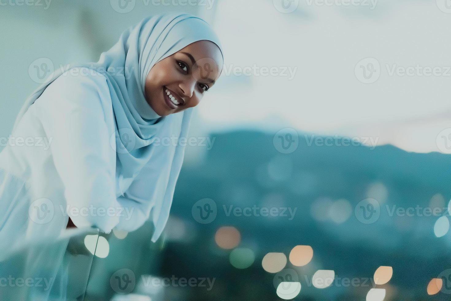 afrikanische muslimische frau in der nacht auf einem balkon lächelt in die kamera mit stadt-bokeh-lichtern im hintergrund. foto