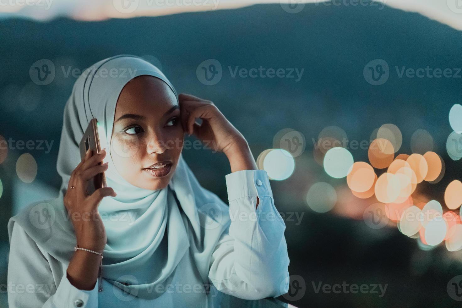 junge muslimische frau mit schalschleier auf der stadtstraße in der nacht, die auf einem smartphone mit bokeh city light im hintergrund sms schreibt. foto