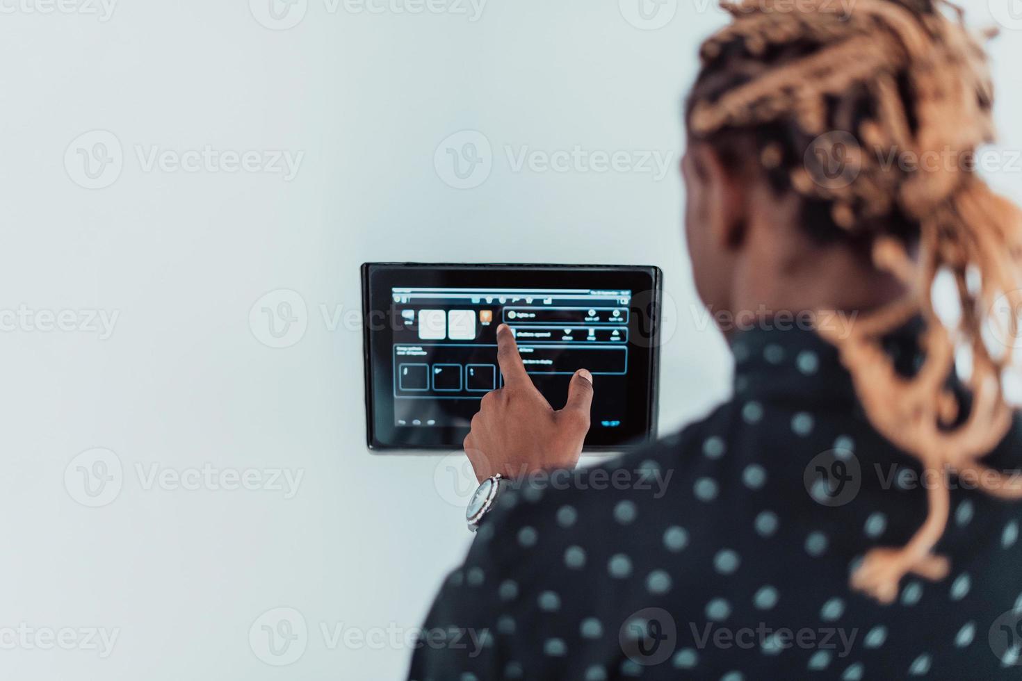 Lächelnder afroamerikanischer Mann mit modernem Smart-Home-System, Controller an der Wand, positiver junger Mann, der die Temperatur am Thermostat schaltet oder Sicherheitsalarm in der Wohnung aktiviert foto