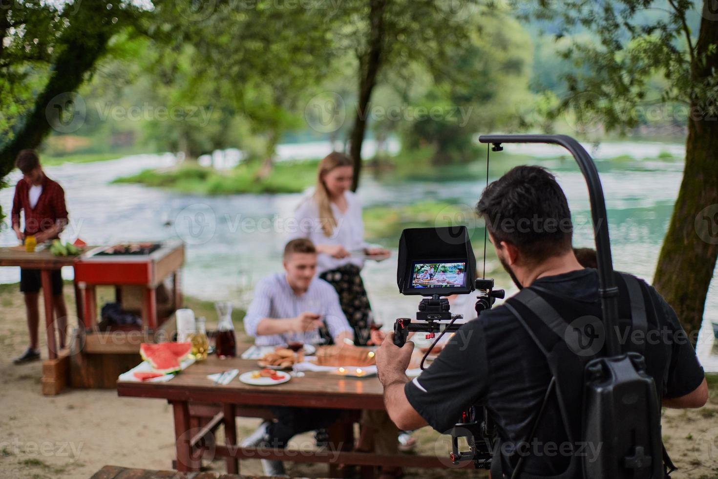 Ein professionell ausgerüsteter Kameramann filmt eine Gruppe von Menschen beim Abendessen am Fluss. foto