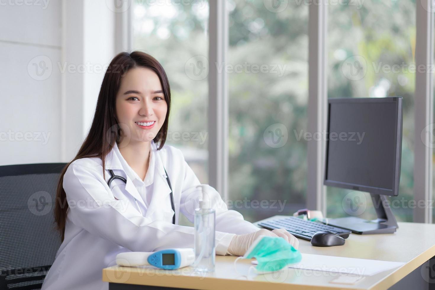 asiatische schöne junge lächelnde ärztin trägt gummihandschuh, der im büro im krankenhaus sitzt. auf dem tisch hat ein papier, eine gesichtsmaske, ein alkoholgel, ein temperaturmessgerät und einen computer. foto