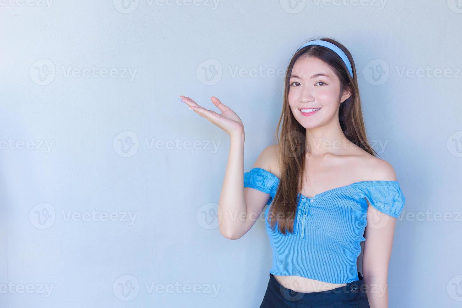 junge schöne asiatische frau in einem blauen hemd fungiert daumen hoch als gutes symbol und eine andere hand zeigt etwas auf weißem hintergrund. foto