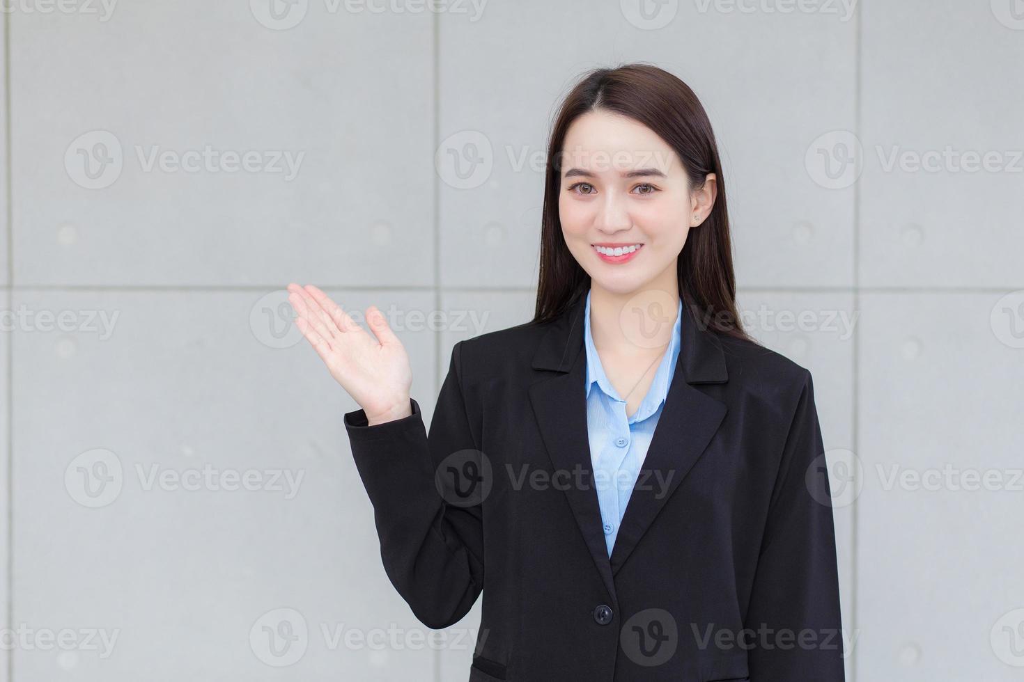 asiatische geschäftsfrau, die einen schwarzen formellen anzug trägt, lächelt und zeigt ihre hand, um etwas an der wand zu präsentieren. foto