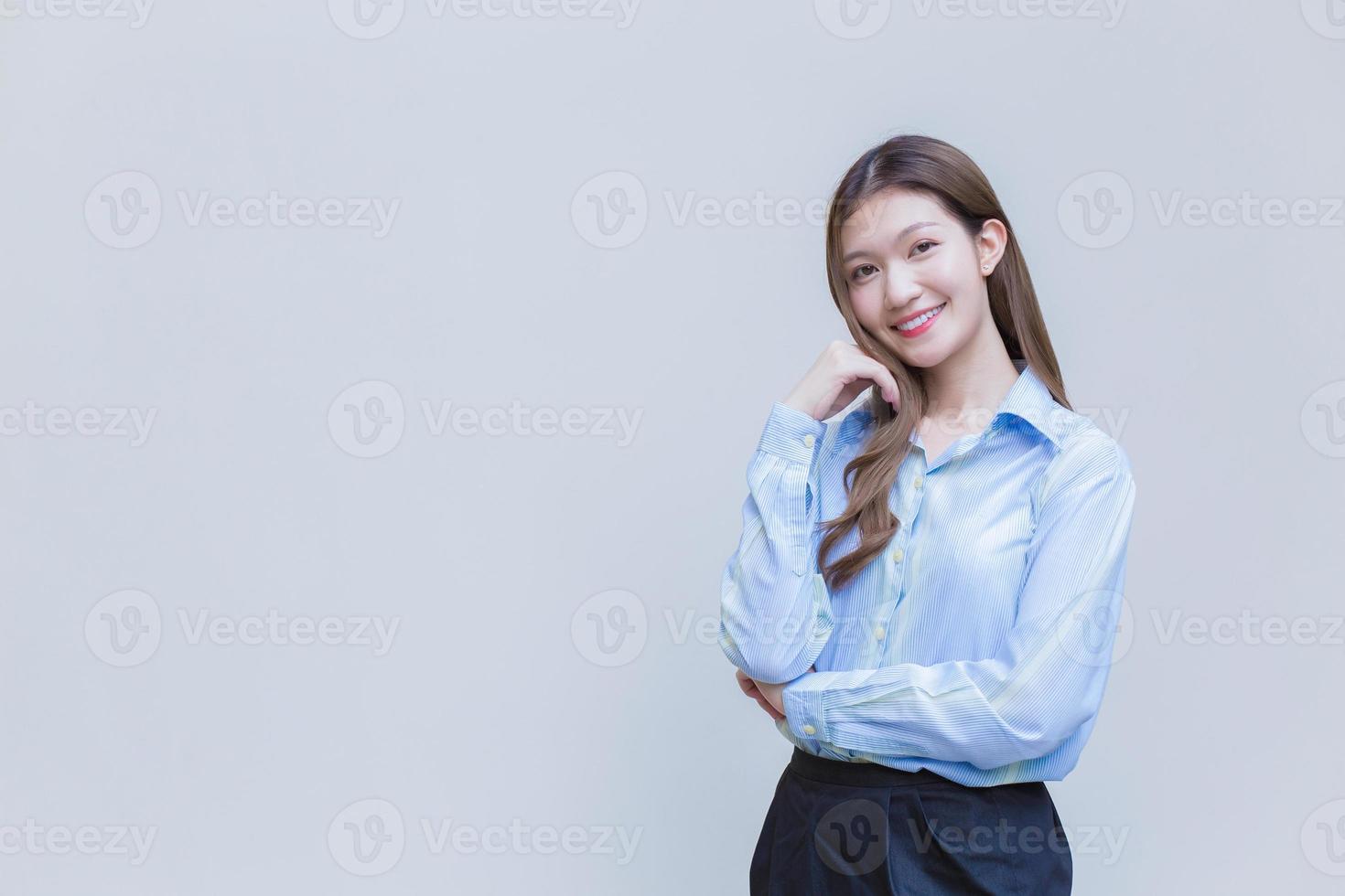 junge asiatische geschäftsfrau mit langen haaren, die ein blaues langarmhemd trägt, lächelt glücklich, während sie die arme verschränkt, um etwas auf weißem hintergrund zu präsentieren. foto