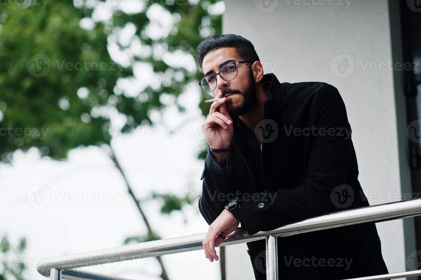 nahöstlicher unternehmer trägt schwarzen mantel und blaues hemd, brille gegen rauchende zigarette des bürogebäudes. foto