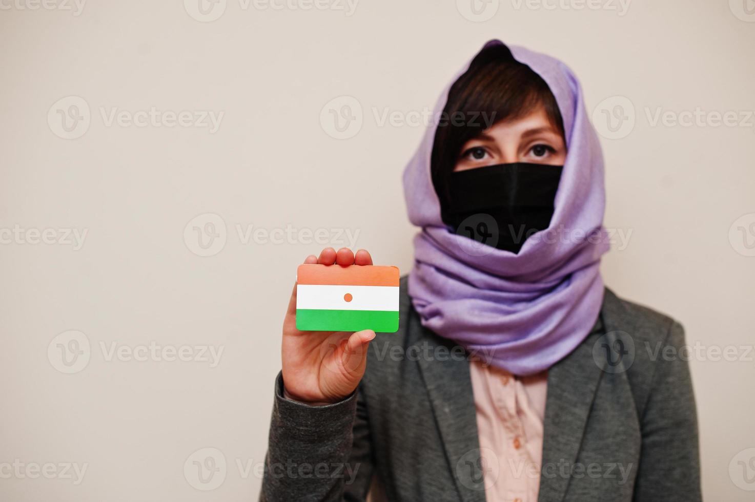 porträt einer jungen muslimischen frau, die formelle kleidung trägt, gesichtsmaske und hijab-kopftuch schützt, niger-flaggenkarte vor isoliertem hintergrund hält. Coronavirus-Länderkonzept. foto