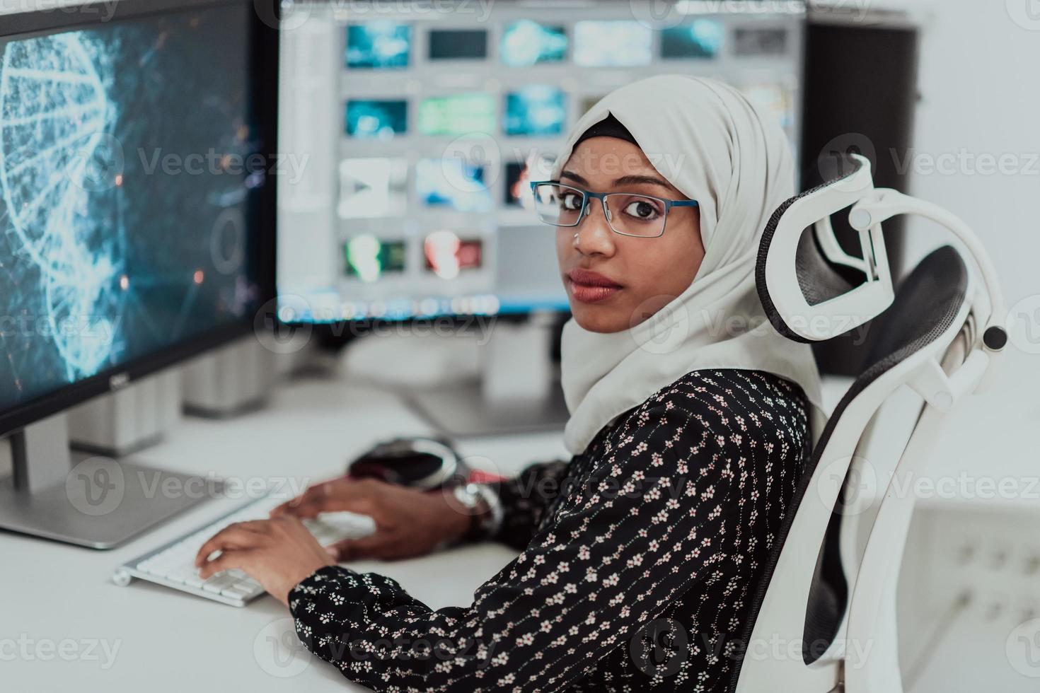 Junge afroamerikanische moderne muslimische Geschäftsfrau mit Schal an einem kreativen, hellen Büroarbeitsplatz mit großem Bildschirm. foto