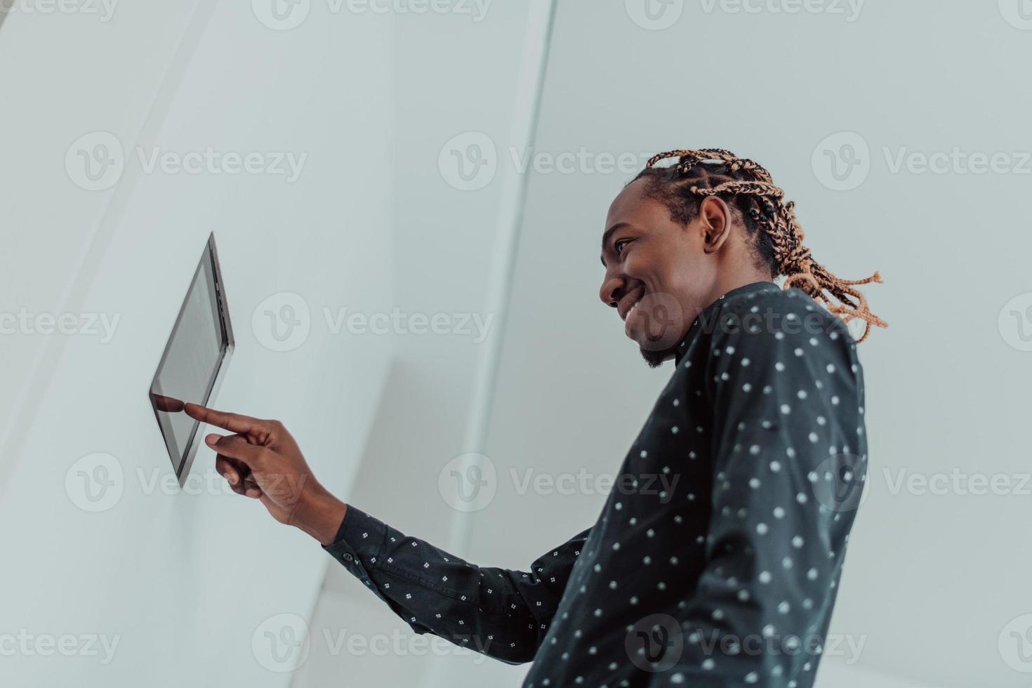 Lächelnder afroamerikanischer Mann mit modernem Smart-Home-System, Controller an der Wand, positiver junger Mann, der die Temperatur am Thermostat schaltet oder Sicherheitsalarm in der Wohnung aktiviert foto