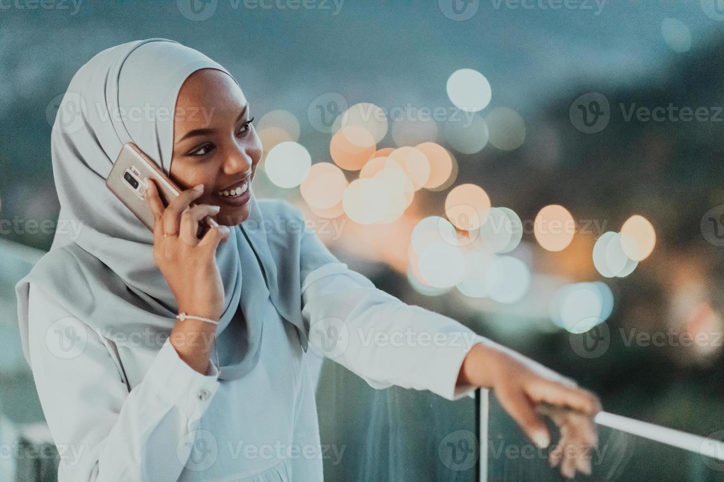 junge muslimische frau mit schalschleier auf der stadtstraße in der nacht, die auf einem smartphone mit bokeh city light im hintergrund sms schreibt. foto
