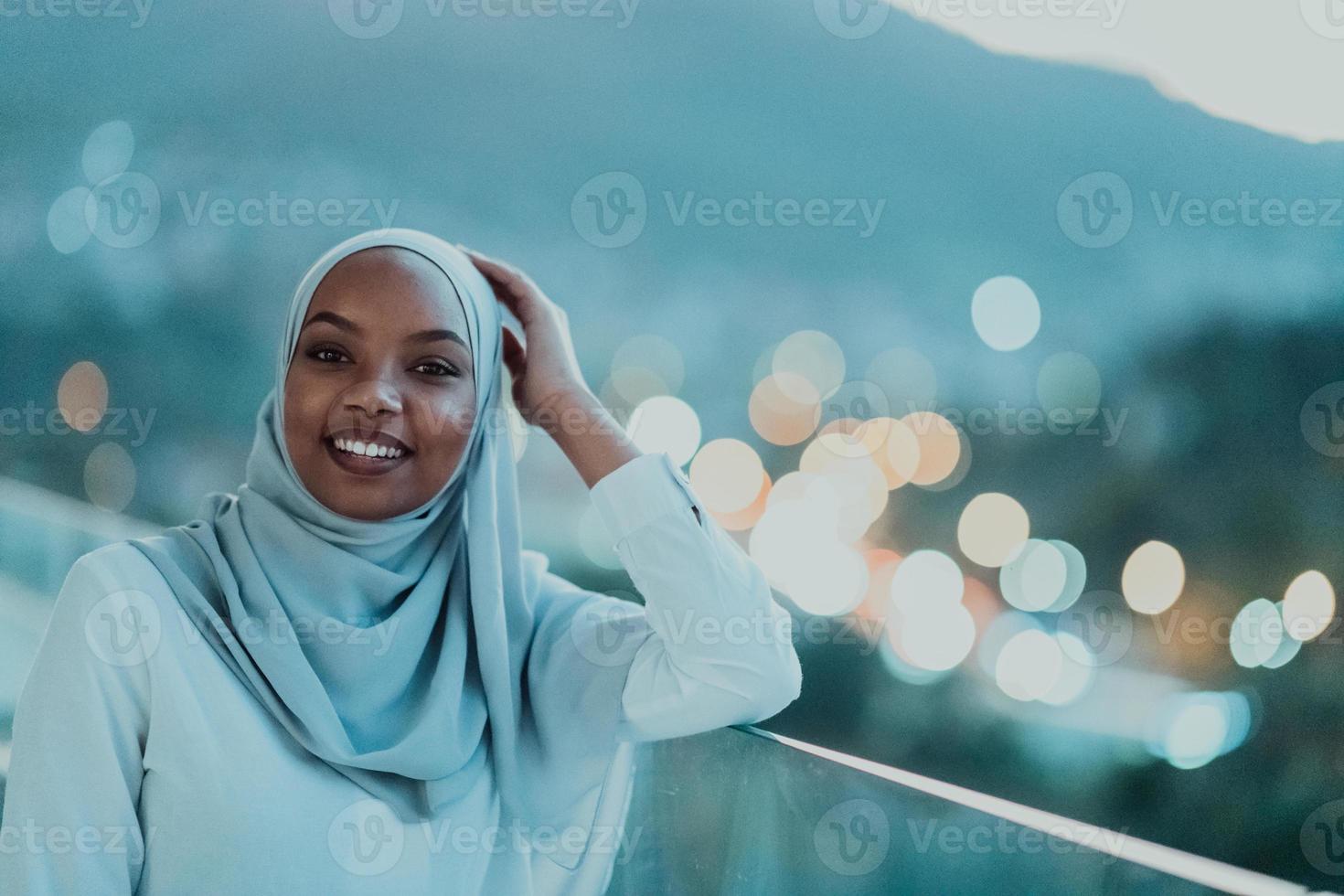 afrikanische muslimische frau in der nacht auf einem balkon lächelt in die kamera mit stadt-bokeh-lichtern im hintergrund. foto