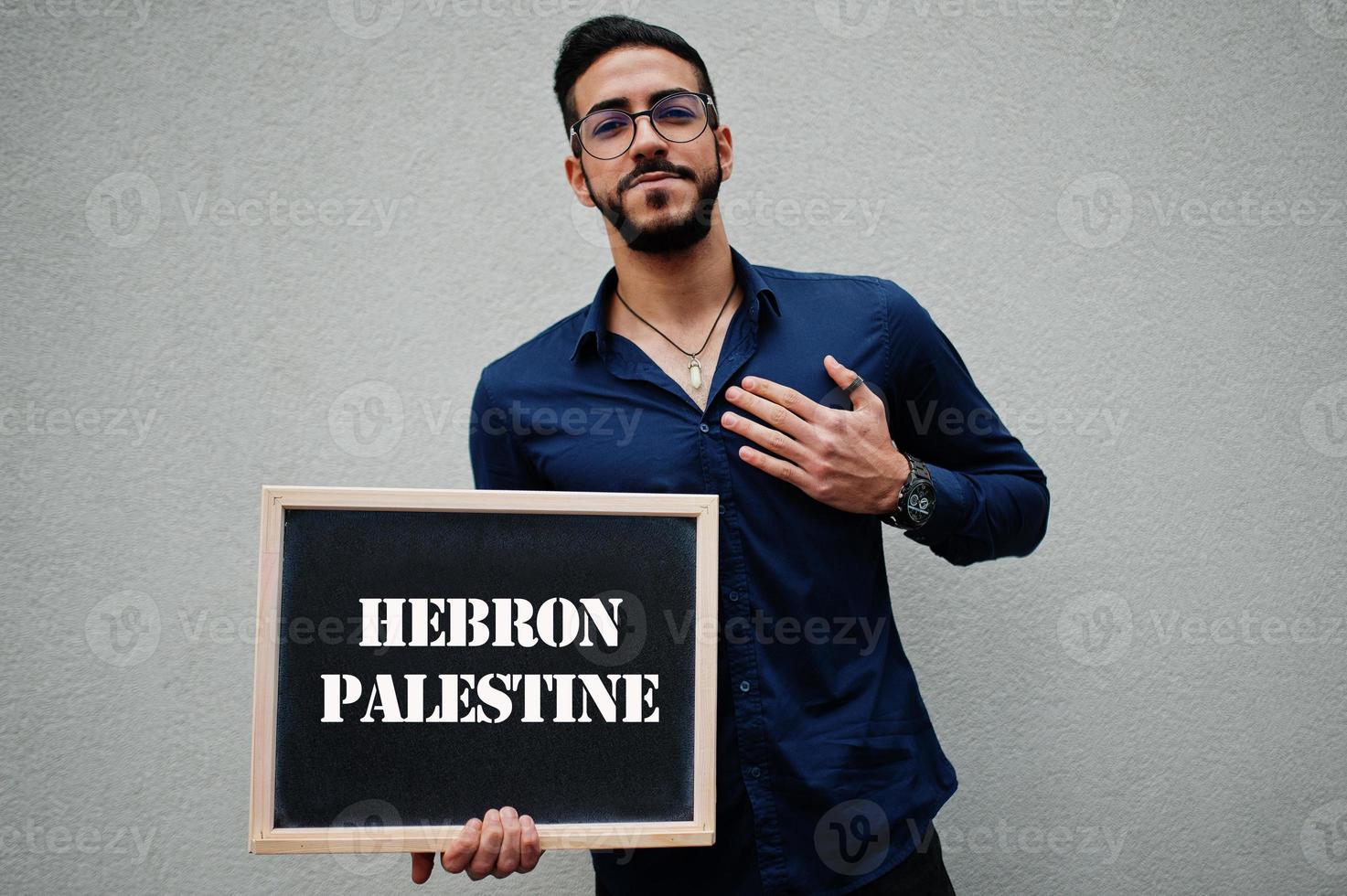 arabischer mann trägt blaues hemd und brille hält tafel mit hebron palästina-aufschrift. größte städte im islamischen weltkonzept. foto