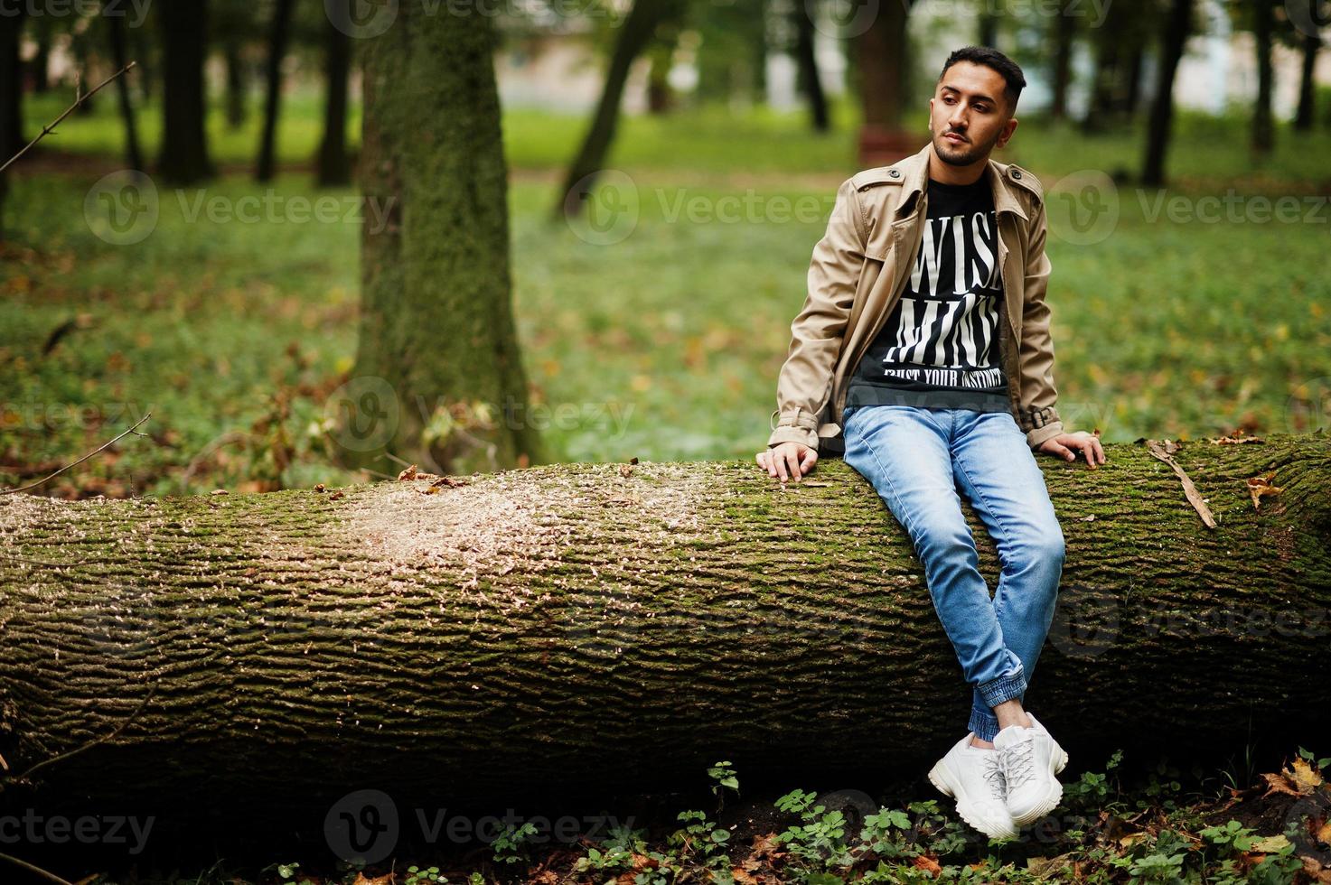 Stilvoller kuwaitischer Mann im Trenchcoat sitzt auf Baum am Holz. foto