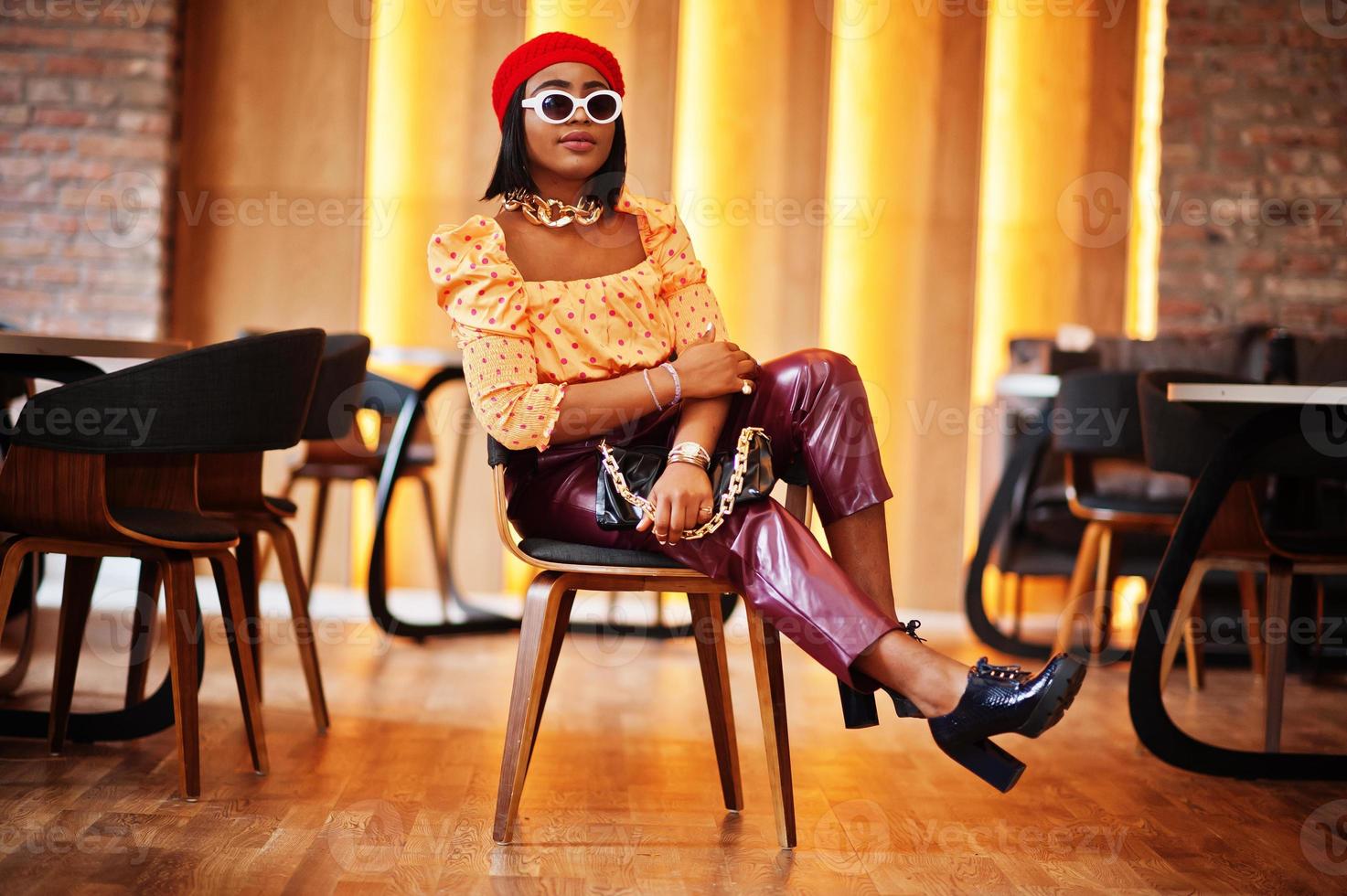 elegante afroamerikanische frau in roter französischer mütze, großer goldener halskette, sonnenbrille, gepunkteter bluse und lederhose posieren drinnen. foto