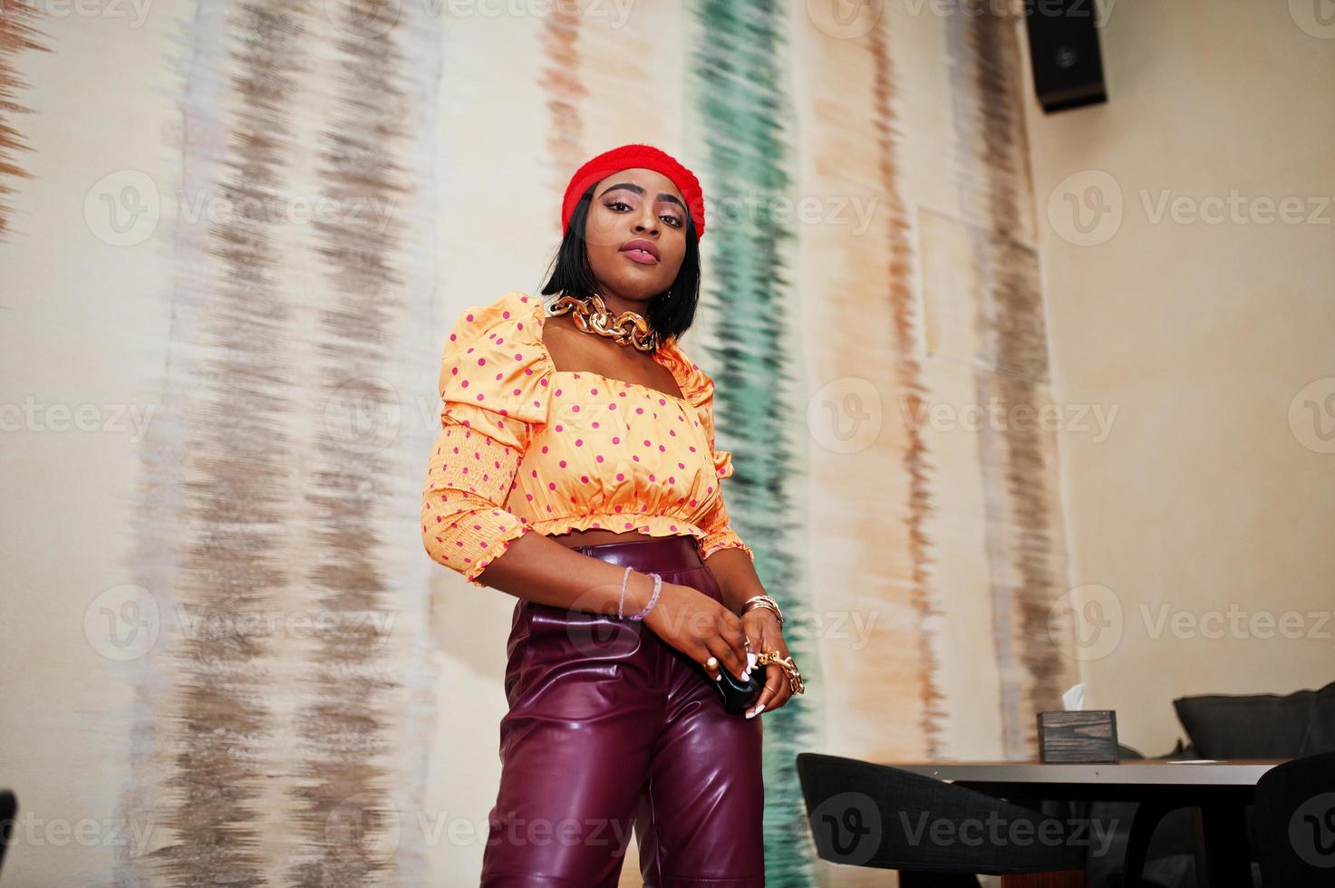 Elegante Afro-Amerikanerin in roter französischer Baskenmütze, großer goldener Halskette, Polka-Dot-Bluse und Lederhose posieren im Innenbereich. foto