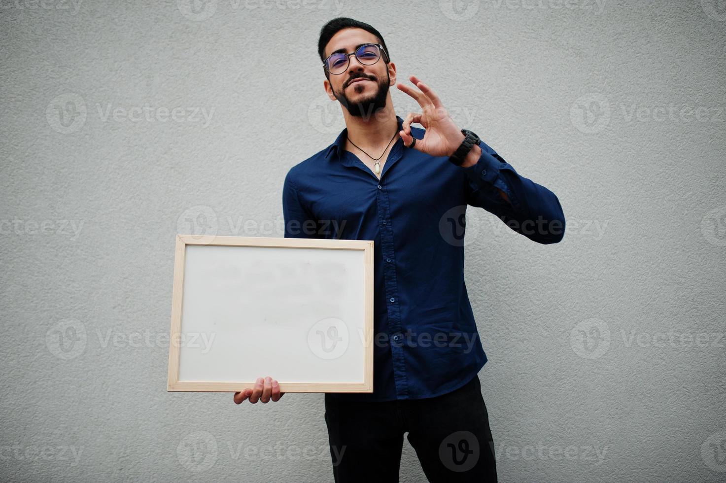 nahöstlicher mann trägt blaues hemd, brille, hält weißes leeres brett. foto