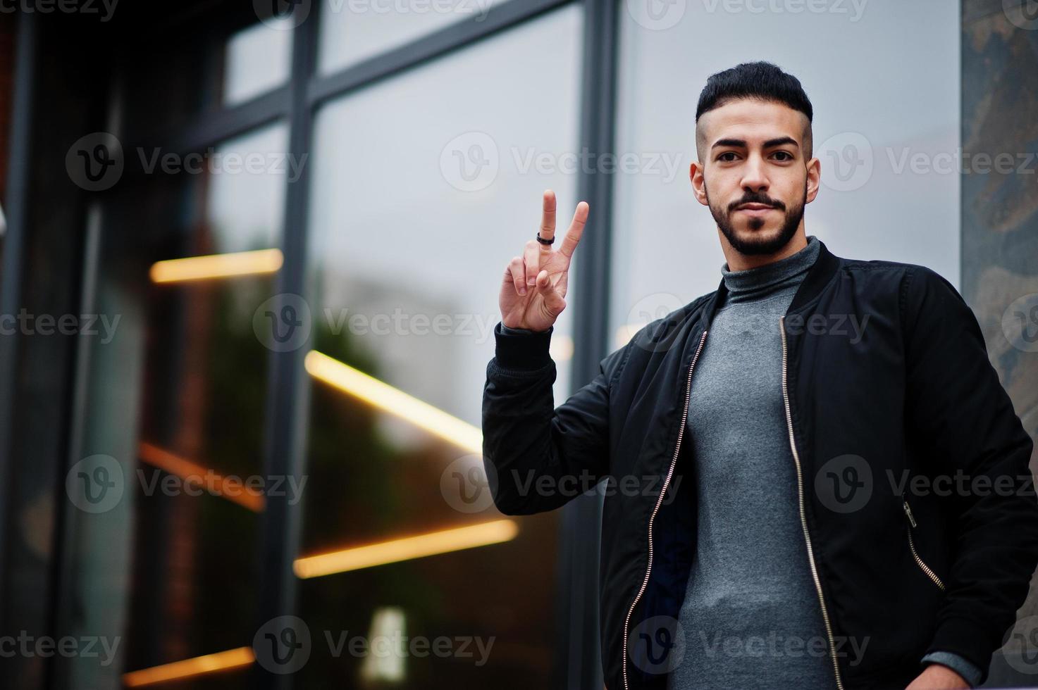 porträt des stilvollen arabischen bartmannes trägt einen grauen rollkragenpullover und eine schwarze jaket. Arabischer Modelltyp zeigt zwei Finger. foto