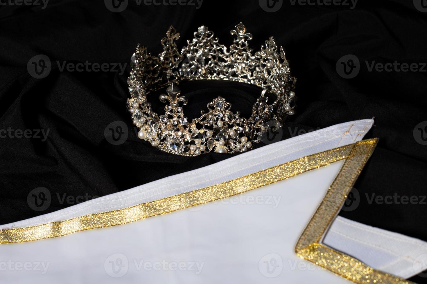 Weißgold-Gewinnerschärpe für Miss-Schönheitswettbewerb, leerer Bereich für Textgewinner-Landwort, Studiobeleuchtung abstrakter dunkler drapierender Textilhintergrund, wichtige Dekoration mit Diamantkrone foto