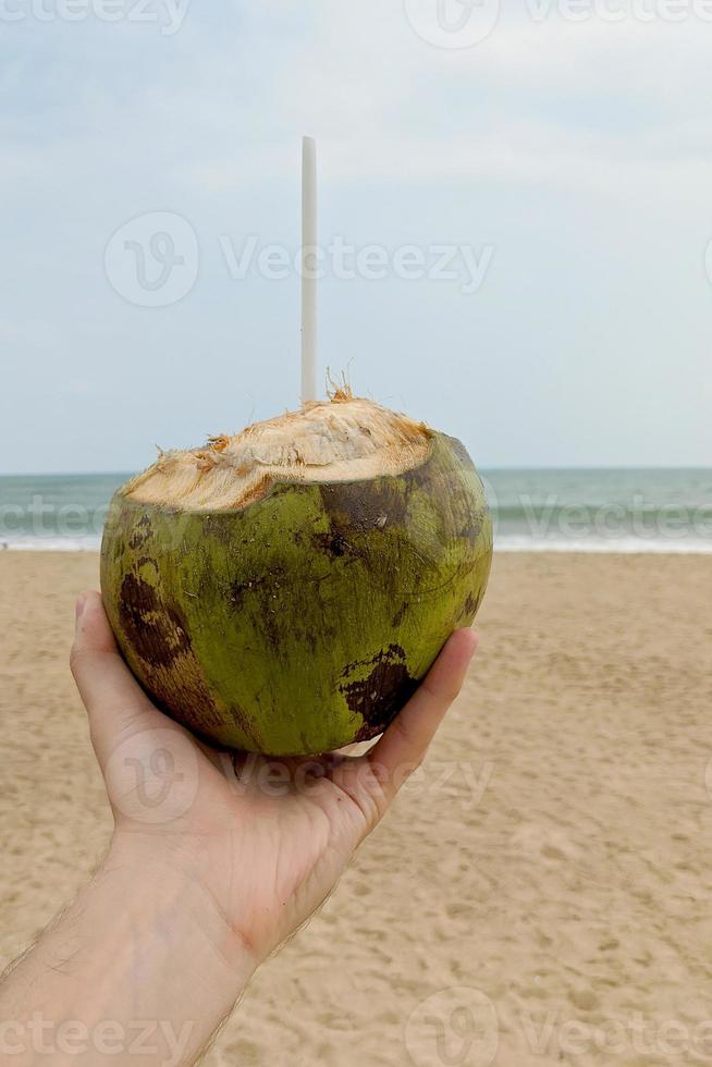 Mann, der reife Kokosnuss mit einem Strohhalm an einem Strand mit blauem Himmel und Wasser im Hintergrund hält foto