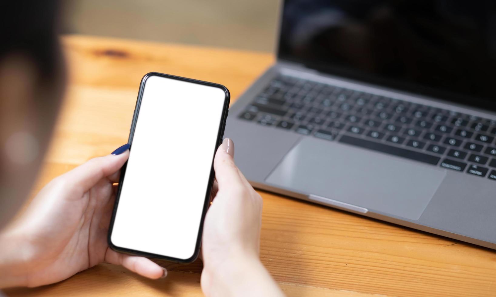 handfrau, die ein telefon, ein leeres bildschirmsmartphone und einen computer auf holztischplatteansicht verwendet. foto