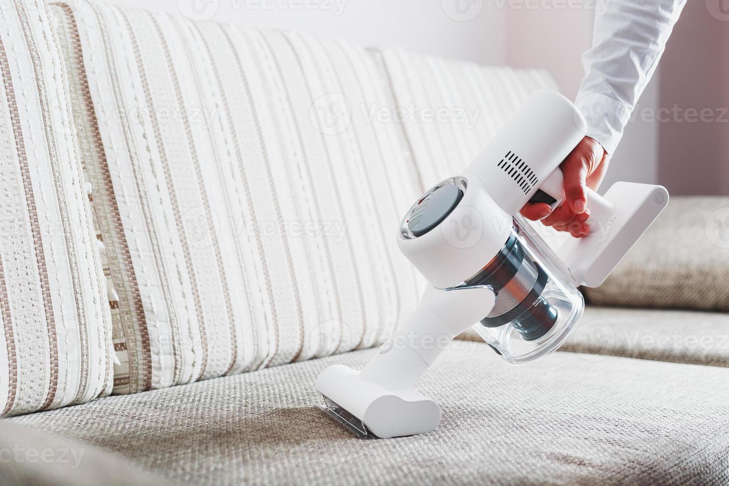 Die Hand eines Mannes hält einen modernen kabellosen Staubsauger zum Reinigen des Sofas im Haus. foto