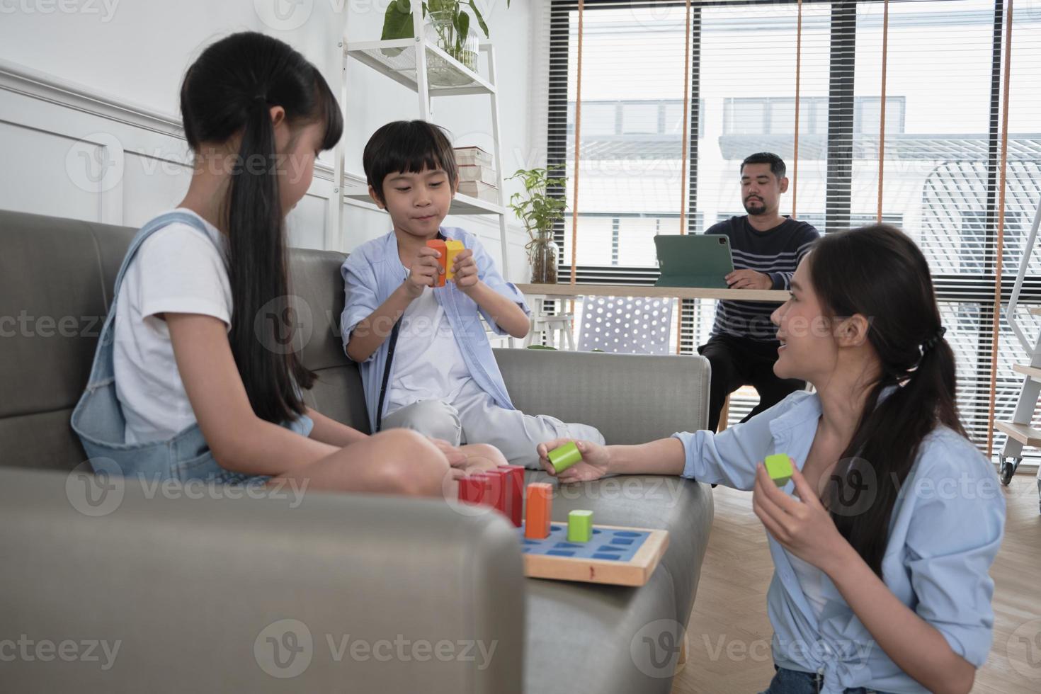 glückliche asiatische thailändische familienpflege, mutter und kleine kinder haben spaß beim gemeinsamen spielen mit bunten spielzeugblöcken auf dem sofa im weißen wohnzimmer, während vater arbeitet, freizeitwochenende und häuslicher wohlfühl-lebensstil. foto