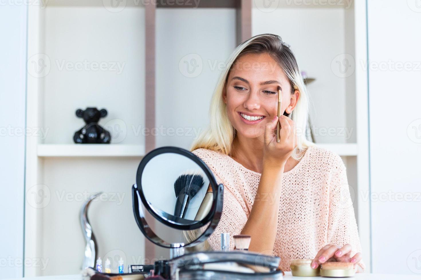 junge schöne frau, die ihr make-up aufträgt und in einen spiegel schaut. schönes Mädchen mit kosmetischem Puderpinsel zum Schminken. bilden. Schminken für perfekte Haut. foto