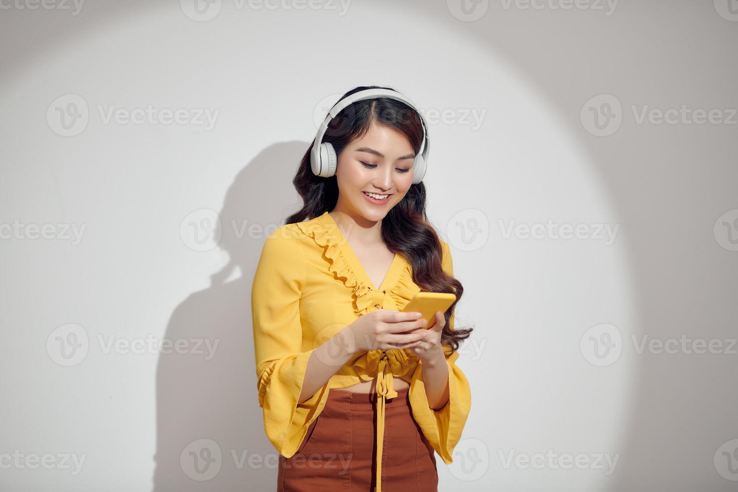 Fröhliche Frau, die Musik mit Kopfhörern isoliert über weißem Hintergrund hört und Handy hält foto