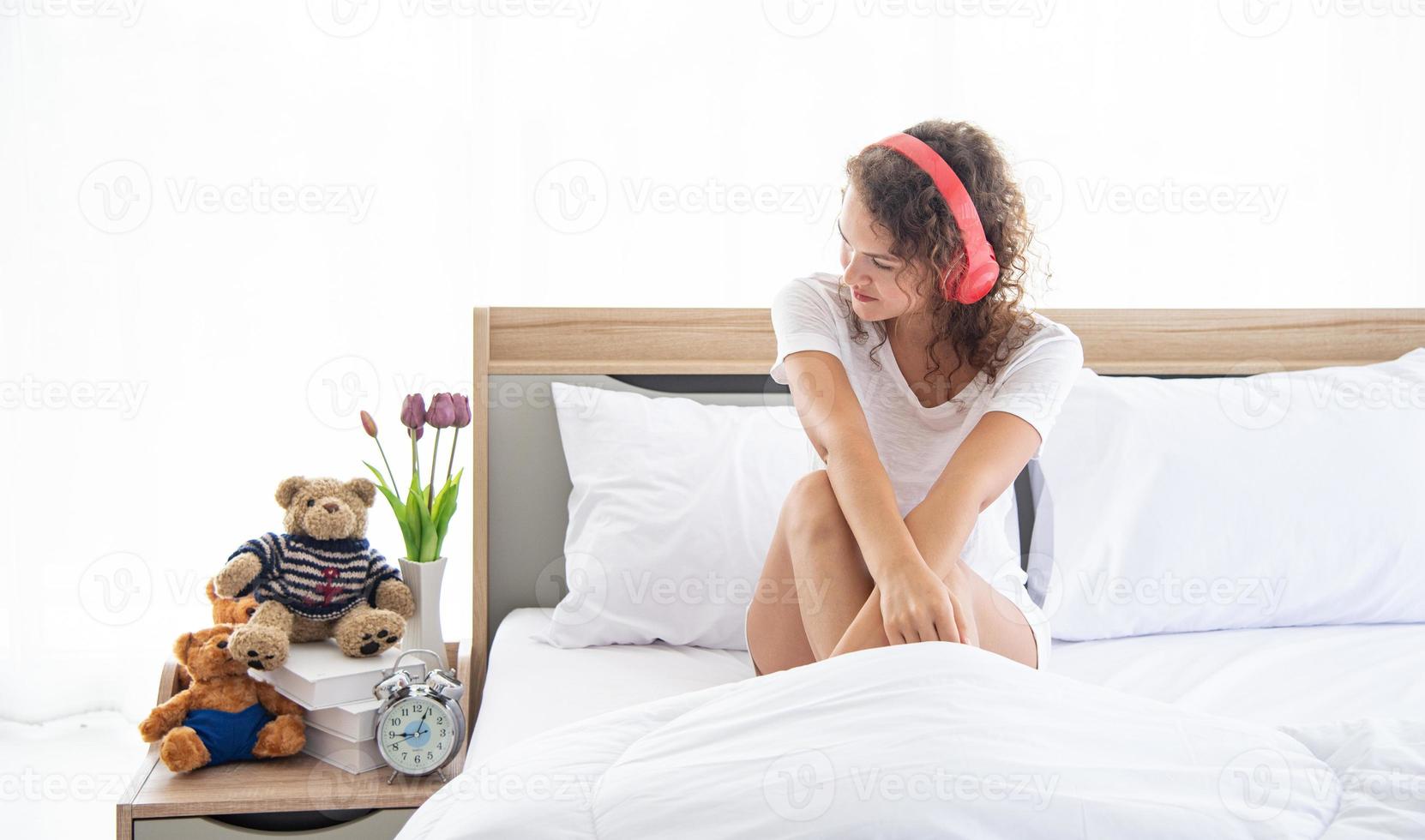 kaukasische junge und schöne glückliche frau mit kopfhörer, die auf dem bequemen doppelbett im modernen schlafzimmer sitzt foto