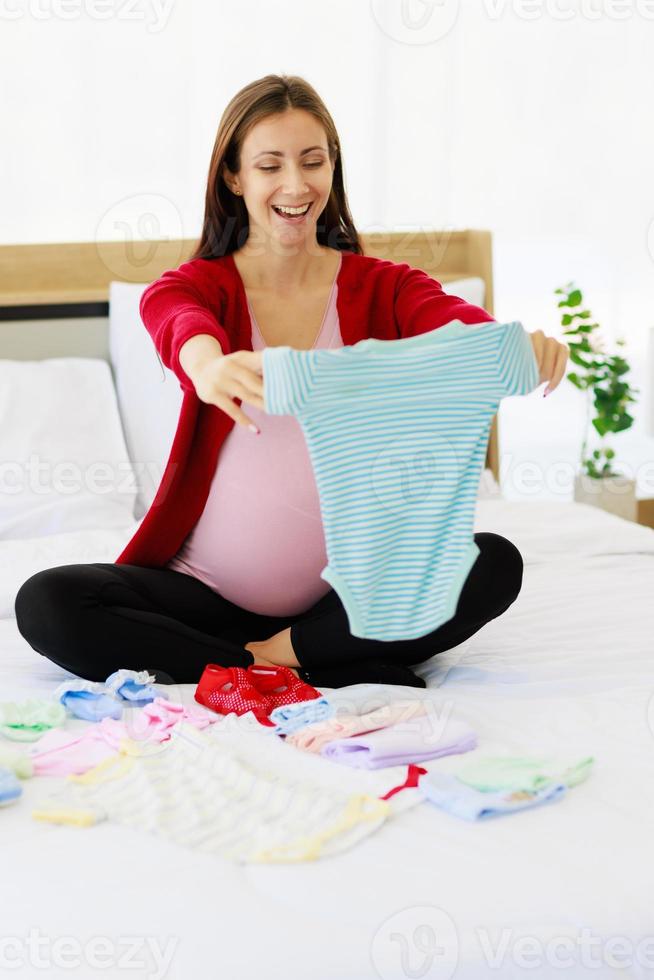 Eine große schwangere Frau bereitet glücklich Babykleidung auf ihrem Bauch vor. foto