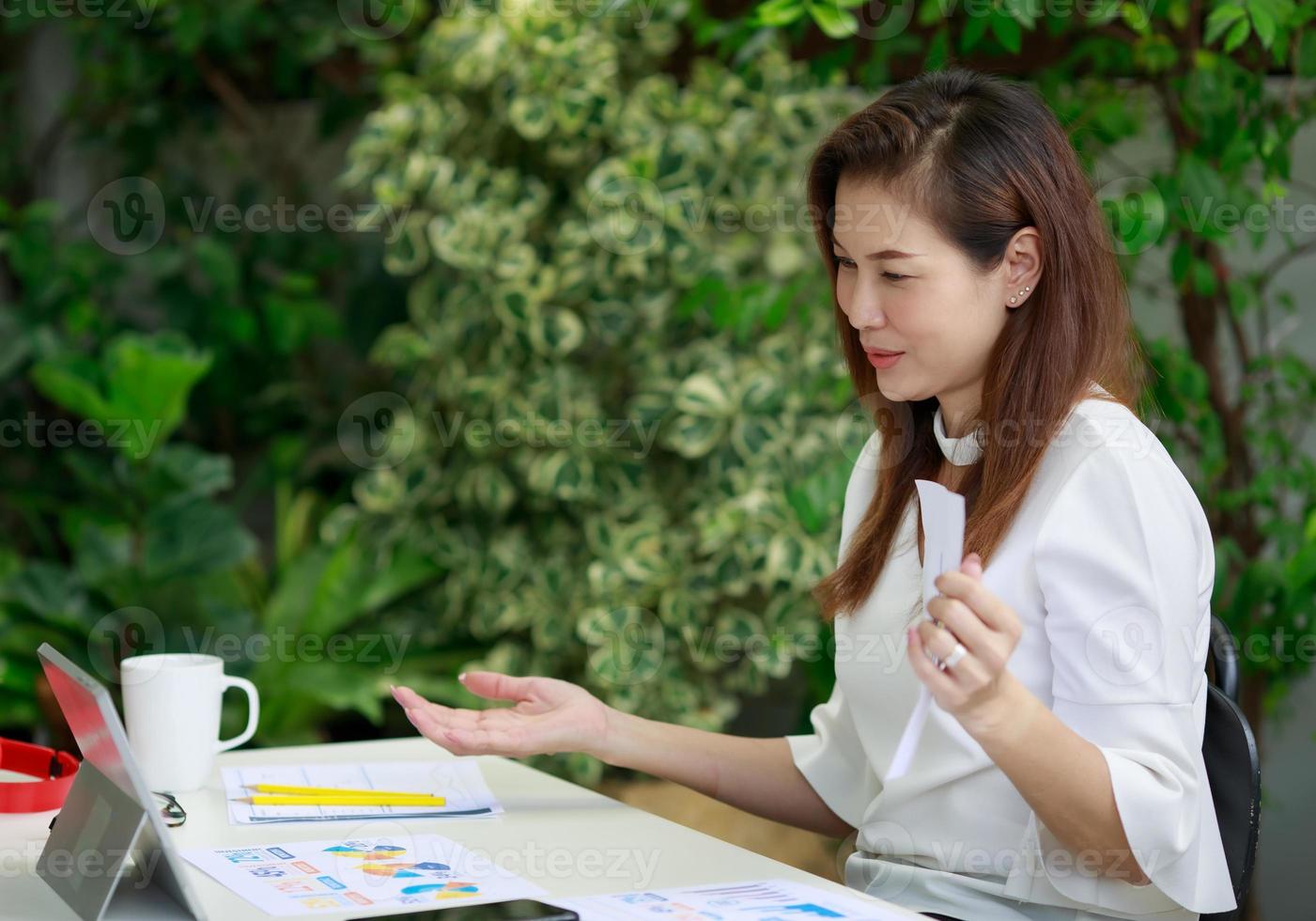 schöne lächelnde asiatische geschäftsfrau, die entspannt im hausgarten sitzt, tablette benutzt und über webanruf kommuniziert, umgeben von exotischen pflanzen. foto