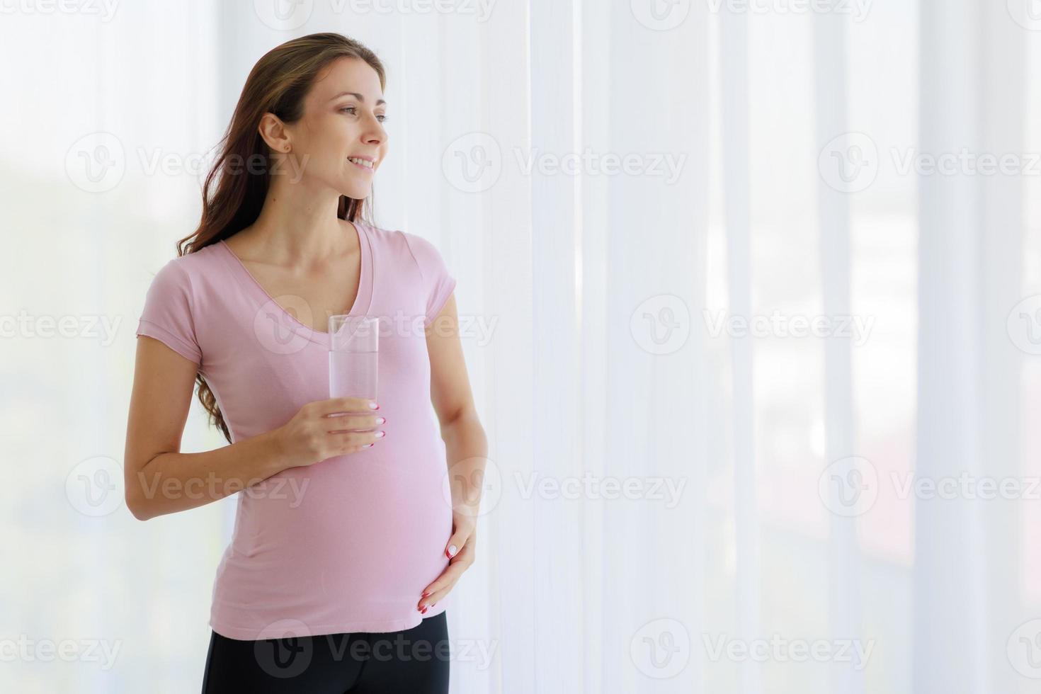 Schöne, fröhliche, schwangere Frau hält ein Glas reines Wasser, um den Körper und das ungeborene Kind zu ernähren. foto