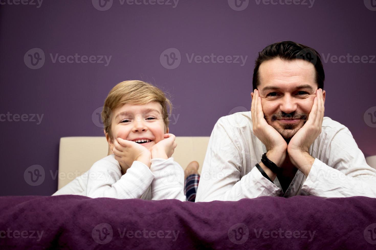 glücklicher sohn und vater, die sich auf das bett legen und in die kamera schauen. foto