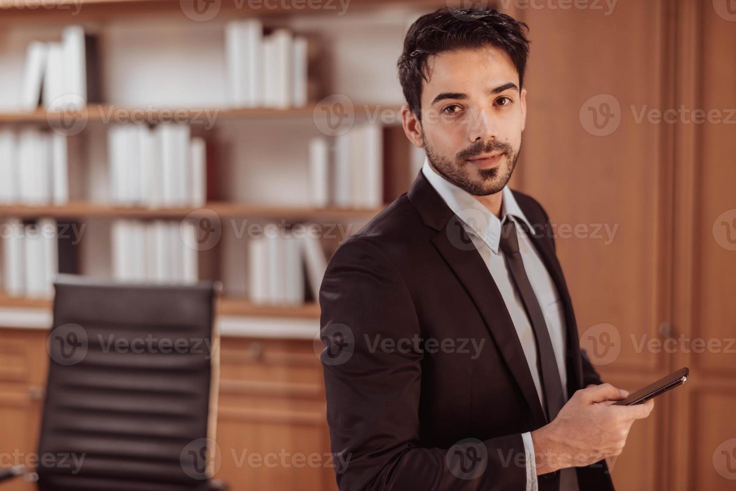 Porträt eines jungen, intelligenten Geschäftsmannes mit Smartphone im Arbeitszimmer. hübscher arabischer geschäftsmann im schwarzen anzug sitzt und hält smartphone für die arbeit. foto