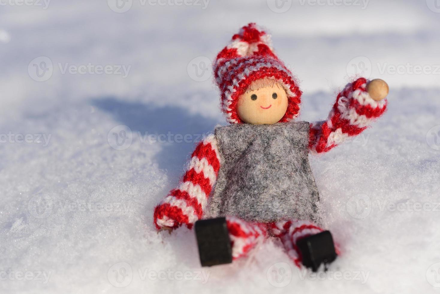 Nahaufnahme einer kleinen Puppe mit Winterkleidung und Hut, die in der Wintersonne im Schnee sitzt foto